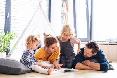 Front view of young family with two small children indoors in bedroom reading a book.